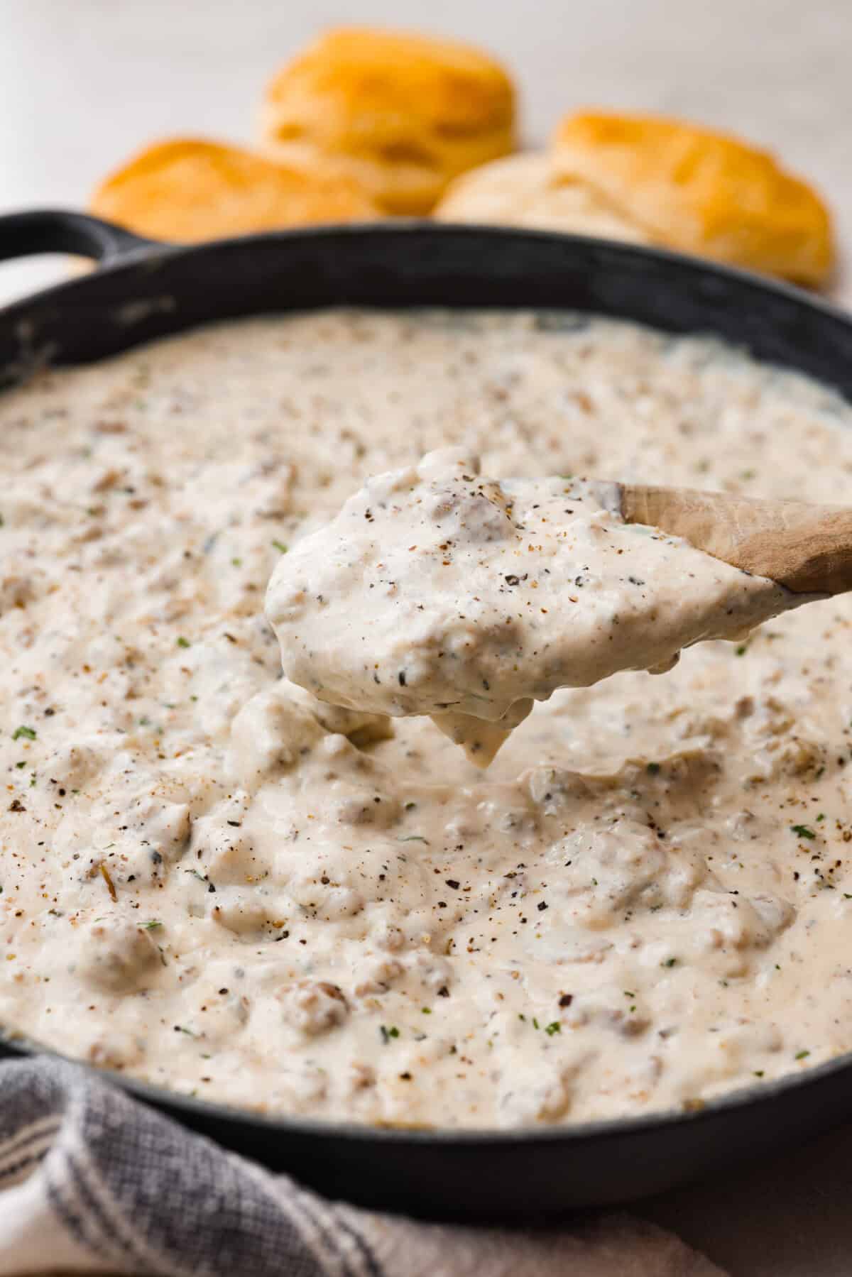 Close up shot of sausage gravy in a skillet with someone scooping some out with a wooden spoon. 